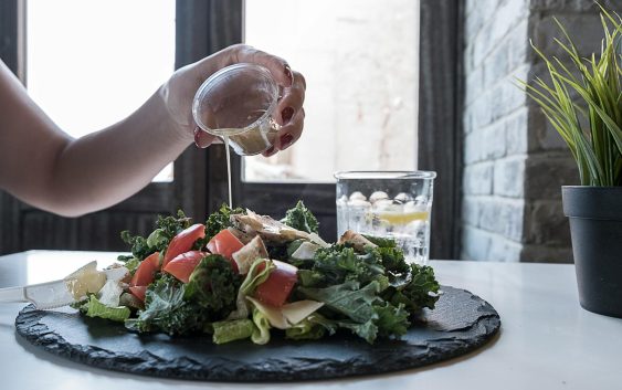 Hand pouring dressing over fresh salad with kale and tomatoes in a modern Dubai restaurant.